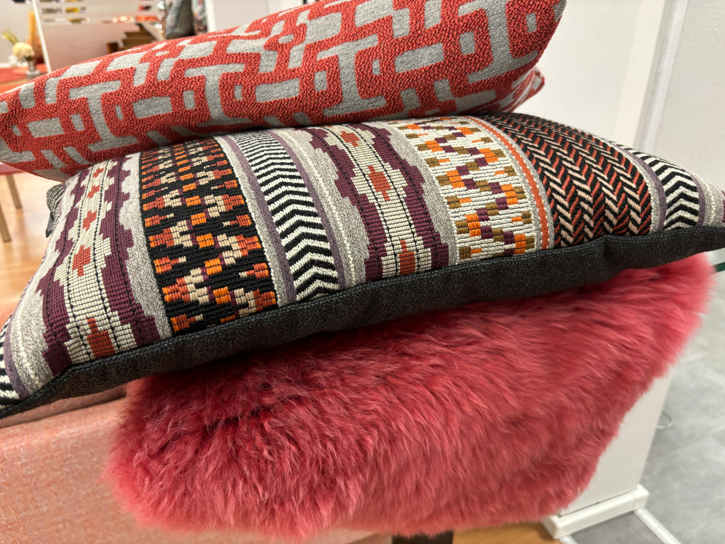 Rohleder Home zwei Kissen. Eines hat ein rotes, geometrisches Muster, das andere ein buntes Streifen- und Zickzackmuster, seitlich von links